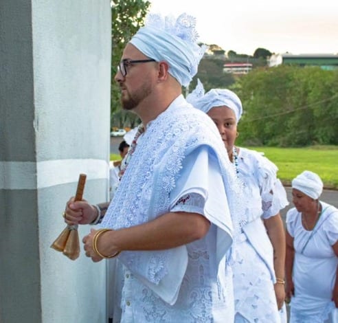 Fotografia de uma pessoa em trajes brancos rendados, com torço branco, óculos e guias de contas ao pescoço, segurando um sino de metal dourado; ao fundo, outras pessoas vestidas de branco, ao ar livre.